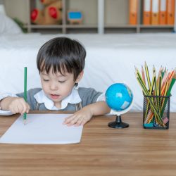 Little boy joyful with use orange pensil color drawing  on white paper, Globe model and Pencil basket on table in the bedroom